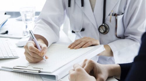 Arzt, der Notizen während einer Patientenberatung auf einem Tisch macht, mit einem Stethoskop um den Hals und einem Computer im Hintergrund.