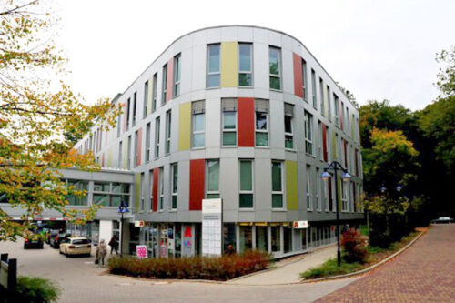 Moderne Architektur eines mehrstöckigen Gebäudes mit bunten Fassadenelementen, umgeben von Bäumen und einem gepflasterten Weg. Ideal für urbane Wohn- und Geschäftsräume.