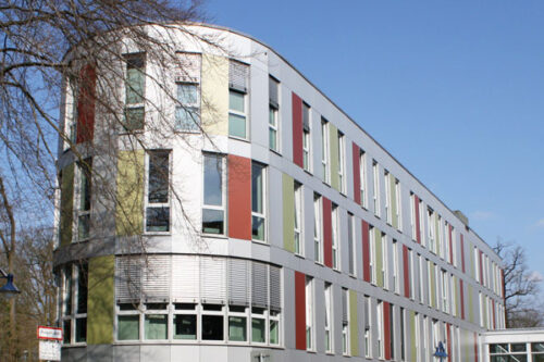 Modernes Gebäude mit farbigen Fassadenelementen und großen Fenstern, umgeben von Bäumen und klarem Himmel. Ideal für Architektur- und Stadtplanungsthemen.