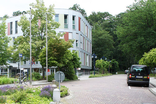 Moderne Wohngebäude an einer ruhigen Straße, umgeben von Bäumen und Grünflächen, mit einem geparkten Auto und einem Fahrradständer im Vordergrund.