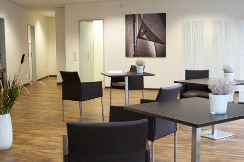 Moderne Büroumgebung mit stilvollen Tischen und Stühlen, Pflanzen und einem dekorativen Wandbild. Helle, einladende Atmosphäre mit Holzfußboden und minimalistischer Einrichtung. Ideal für Meetings oder entspannte Gespräche.