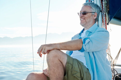 Ein älterer Mann mit Brille sitzt entspannt auf einem Segelboot und genießt die Aussicht auf das Wasser und die Berge im Hintergrund. Er trägt ein hellblaues Hemd und khakifarbene Shorts.