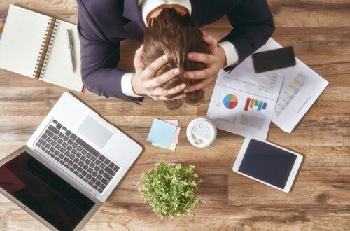 Mann in Anzug mit gestresstem Gesichtsausdruck sitzt an einem Schreibtisch, umgeben von Laptop, Smartphone, Tablet, Notizblock und Dokumenten mit Diagrammen und Statistiken.