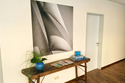 Ein moderner Flur mit einem Holzschreibtisch, auf dem Bücher und Zeitschriften liegen. Im Hintergrund hängt ein großes schwarz-weißes Bild von Segeln. Eine grüne Pflanze in einem blauen Topf ergänzt die Einrichtung. Die Wand ist hell gestrichen, und eine Tür führt in einen weiteren Raum.