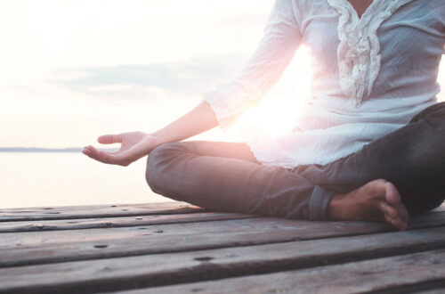 Person in Meditationshaltung auf einem Holzsteg am Wasser, umgeben von sanftem Licht und einer ruhigen Atmosphäre.