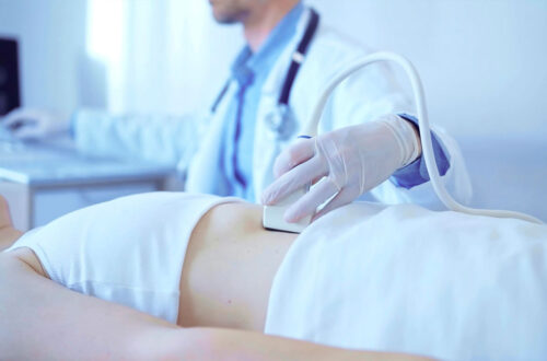 Ein Arzt führt eine Ultraschalluntersuchung am Bauch eines Patienten durch. Der врач trägt Handschuhe und verwendet ein Ultraschallgerät, während im Hintergrund medizinische Ausrüstung sichtbar ist.