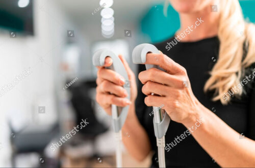 Frau hält zwei Geräte in den Händen, die für eine physiotherapeutische oder kosmetische Behandlung verwendet werden. Unscharfer Hintergrund zeigt eine moderne Praxisumgebung.