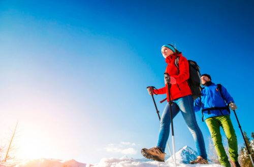 Winterwanderung in den Bergen: Ein Mann und eine Frau in warmer Kleidung mit Wanderstöcken gehen durch den Schnee an einem sonnigen Tag.