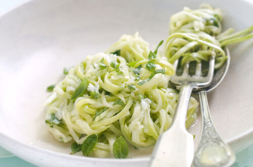 Zucchini-Nudeln mit frischem Basilikum und essbaren Blüten in einer weißen Schüssel, serviert mit einer Gabel und einem Löffel. Ideal für eine gesunde, vegetarische Mahlzeit.