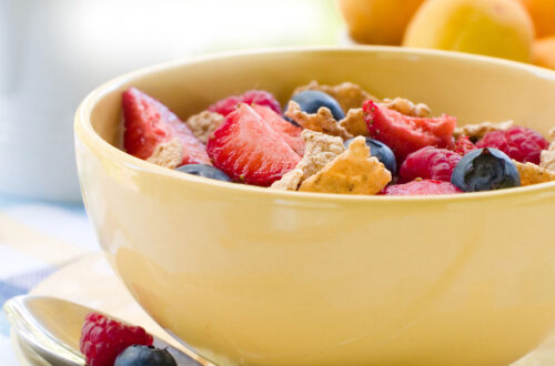 Schüssel mit Müsli, frischen Erdbeeren, Himbeeren und Heidelbeeren, serviert auf einem Tisch mit frischem Obst im Hintergrund. Ideal für ein gesundes Frühstück.