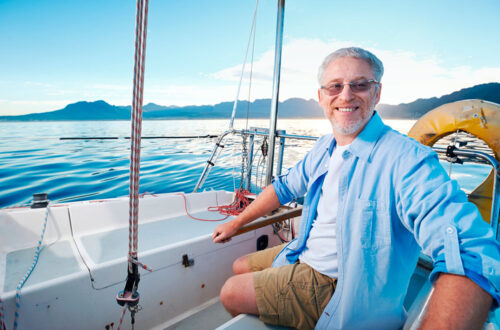 Ein lächelnder älterer Mann sitzt auf einem Segelboot mit Blick auf ruhiges Wasser und Berge im Hintergrund. Er trägt ein helles Hemd und Shorts, ideal für einen perfekten Tag auf dem Wasser.