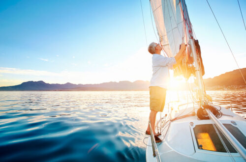 Ein älterer Mann, der auf einem Segelboot steht und das Segel bei Sonnenuntergang hochzieht. Im Hintergrund sind Berge und ruhige Wasseroberfläche zu sehen.