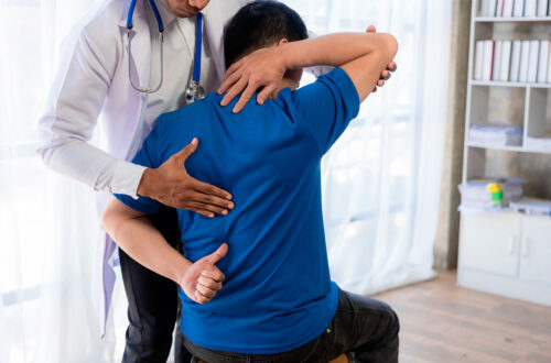 Arzt untersucht einen Patienten mit Rückenschmerzen, während er eine therapeutische Technik anwendet. Der Patient sitzt und trägt ein blaues T-Shirt, während der Arzt ihm mit einer Hand an den Rücken fasst und mit der anderen dessen Arm unterstützt. Hinter ihnen ist ein hell erleuchteter Raum mit einer Regaleinheit im Hintergrund.