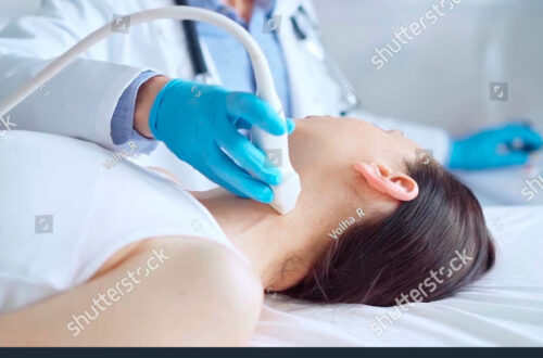 Ärztliche Untersuchung einer Patientin mit einem Ultraschallgerät am Hals, das für diagnostische Zwecke eingesetzt wird. Im Hintergrund ist ein Arzt in einem weißen Kittel zu sehen.