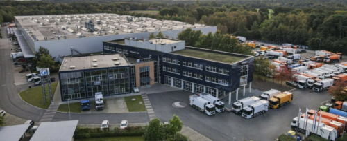 Luftaufnahme eines modernen Logistikzentrums mit mehreren Lkw auf dem Parkplatz und einem großen Gebäude im Vordergrund. Im Hintergrund sind Lagerhallen und Bäume zu sehen.