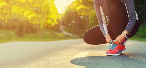 Frau in Sportbekleidung bindet ihre Schuhe auf einem sonnigen Waldweg, ideal für Jogging oder Outdoor-Aktivitäten.