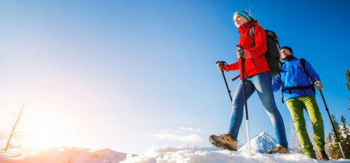 Zwei Personen beim Schneewandern in den Bergen mit klar blauem Himmel und sonnigem Wetter. Die Frau trägt eine rote Jacke und die Männer eine blaue Jacke. Beide nutzen Wanderstöcke und bewegen sich durch frischen Schnee.